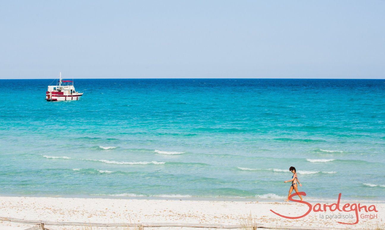 A woman walks on the white sand beach of La Cinta with the light blue sea with a boat in the background