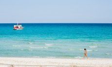 Porto Taverna - Caribbean flair | A woman walks on the white sand beach of La Cinta with the light blue sea with a boat in the background A woman walks on the white sand beach of La Cinta with the light blue sea with a boat in the background